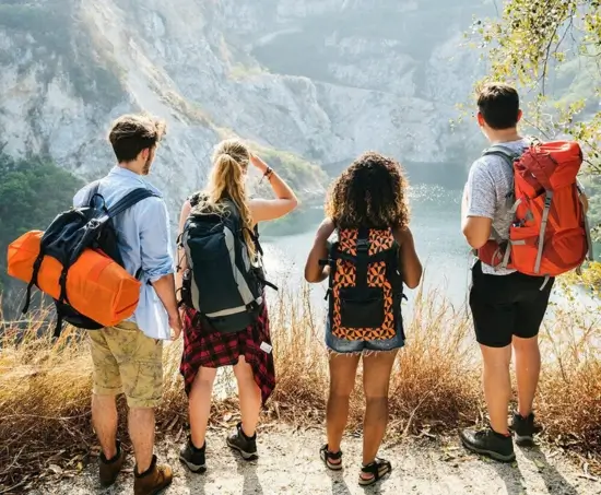 Four friends with hiking backpacks stand at the edge of a trail, overlooking a serene mountain lake in Malaysia's rugged highlands, ready for an adventure with MJ Adventure Travel Services.