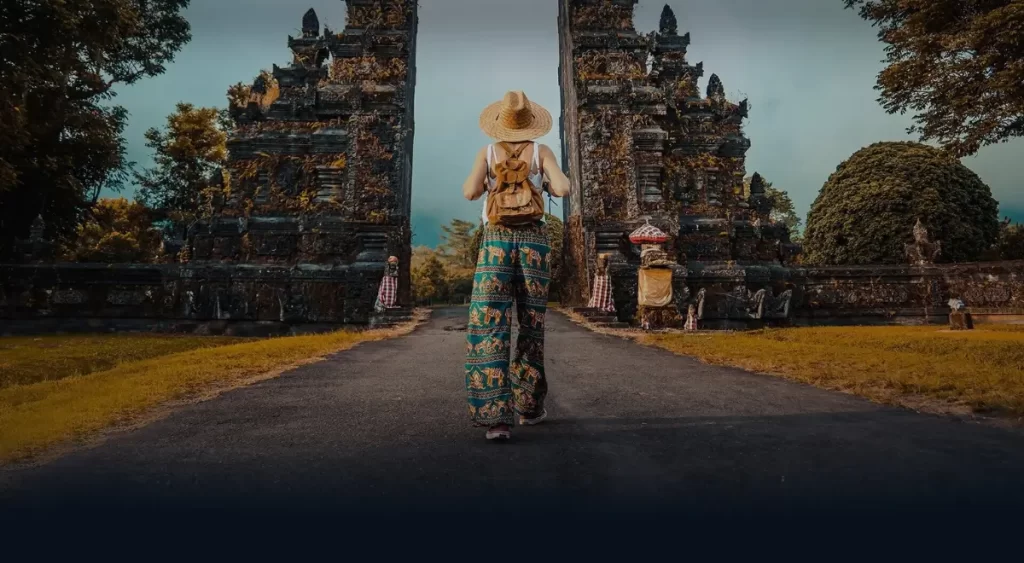 A solo traveler dressed in colorful attire walks towards the towering, intricately carved gates of a Balinese temple, capturing the essence of spiritual and cultural exploration in Bali.